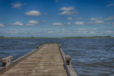 Old rickety wooden jetty leading into a calm lake reflecting the sunny blue sky with green shoreline and trees in the distanceの写真素材