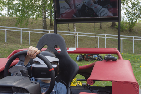Child wearing VR glasses driving a red car in a playground turning the wheel as though cornering as he reacts with his virtual environmentの写真素材