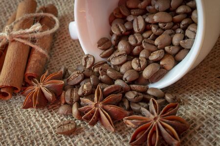 Roasted coffee beans spilling from a cup, cinnamon with star anise spice on a on jute sack in a close up viewの写真素材