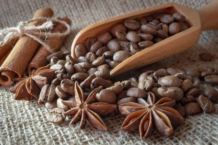 Roasted coffee beans spilling from a wooden scoop, cinnamon sticks and star anise spice on a on jute sack in a close up viewの写真素材
