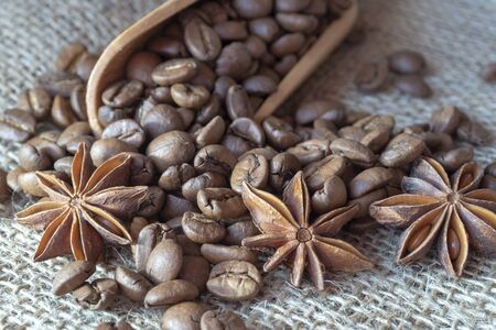 Roasted coffee beans spilling from a wooden scoop, cinnamon sticks and star anise spice on a on jute sack in a close up viewの写真素材
