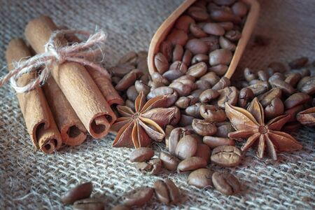 Roasted coffee beans spilling from a wooden scoop, cinnamon sticks and star anise spice on a on jute sack in a close up viewの写真素材