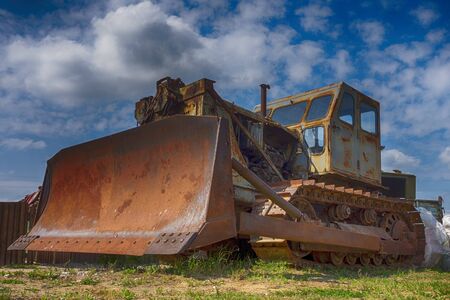 Low angle view of an old rusty heavy duty industrial digger or grader parked outdoors in a field under a cloudy blue skyの写真素材
