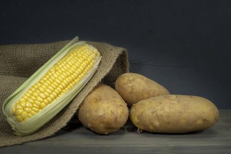 Raw, unpeeled potatoes and corncobs spilling out of hessian sack on a rustic old wooden tabletop, black wall in background, low angle viewの写真素材