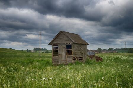 Old dilapidated wooden hut in a farm field in spring with lush green grass under a threatening sky with grey cloudsの写真素材