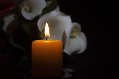 A close up of an orange candle and flame and lily flowers on a dark background.の写真素材