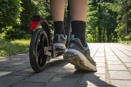 Teenager riding black kick scooter on road in park at summer sunny day, close-up rear horizontal viewの写真素材