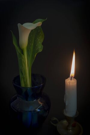 A close up of an orange candle and flame and lily flowers on a dark background.の写真素材
