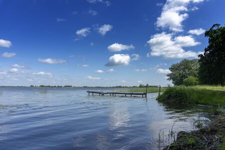 Old rickety wooden jetty leading into a calm lake reflecting the sunny blue sky with green shoreline and trees in the distanceの写真素材