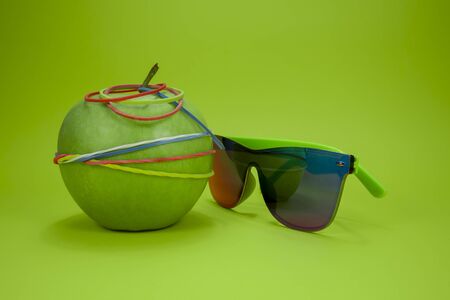 Green apple tied in colorful rubber bands and fashion sunglasses over a green background. Simplicity back to school or diet, aerobic concept with copy spaceの写真素材