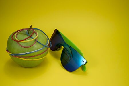 Green apple tied in colorful rubber bands and fashion sunglasses over a yellow background. Simplicity back to school or diet, aerobic concept with copy spaceの写真素材