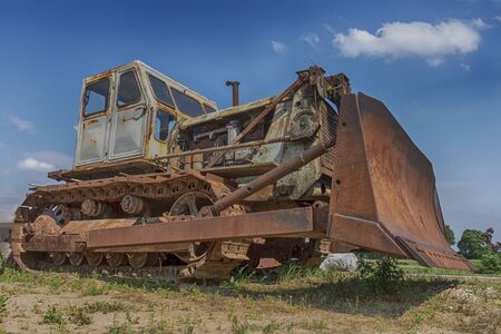 Low angle view of an old rusty heavy duty industrial digger or grader parked outdoors in a field under a cloudy blue skyの写真素材