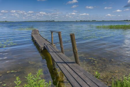 Old rickety wooden jetty leading into a calm lake reflecting the sunny blue sky with green shoreline and trees in the distanceの写真素材