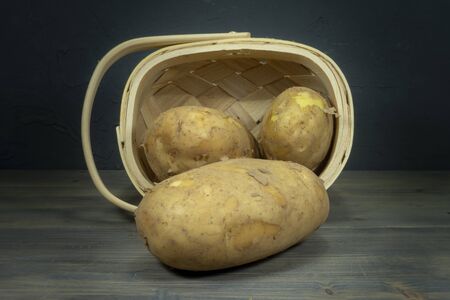 Fresh potatoes spilling out of tipped wicker basket onto grey wooden tabletop. Black wall in background, low angle viewの写真素材