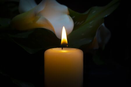 A close up of an orange candle and flame and lily flowers on a dark background.の写真素材