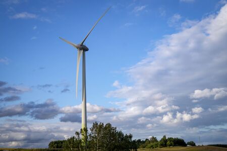 Wind power turbines for electric power generation in landscape with green grass, trees and cloudy blue skyの写真素材