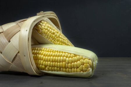 Fresh sweetcorn cobs spilling out of tipped wicker basket onto grey wooden tabletop. Black wall in background, low angle viewの写真素材