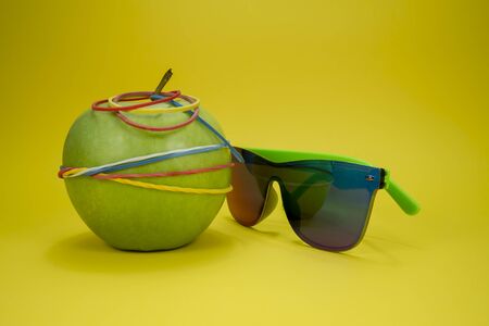 Green apple tied in colorful rubber bands and fashion sunglasses over a yellow background. Simplicity back to school or diet, aerobic concept with copy spaceの写真素材