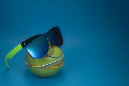 Green apple tied in colorful rubber bands and fashion sunglasses over a blue background. Simplicity back to school or diet, aerobic concept with copy spaceの写真素材