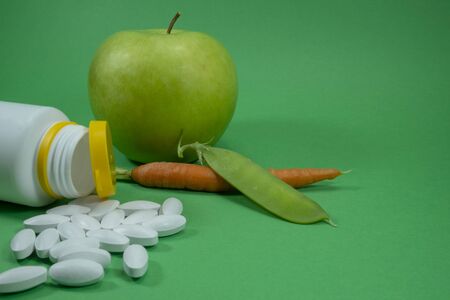 Pills spilling out of a white bottle near apple, pea pod and carrot on a green background. Healthcare, diet or weight loss conceptの写真素材