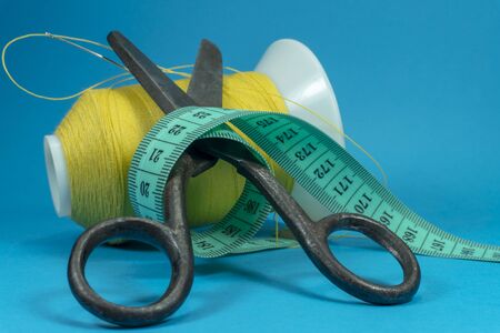 Old metal scissors wrapped around with tape measure and spools of thread on blue background, needlework and tailoring concept with free copy spaceの写真素材