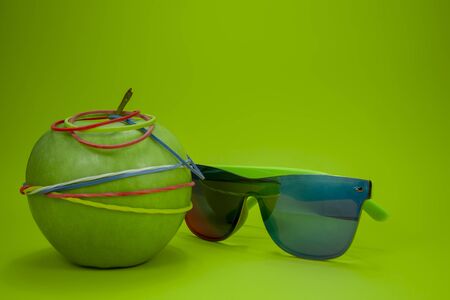 Green apple tied in colorful rubber bands and fashion sunglasses over a green background. Simplicity back to school or diet, aerobic concept with copy spaceの写真素材