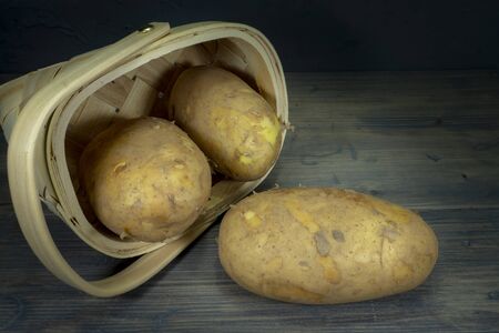 Fresh potatoes spilling out of tipped wicker basket onto grey wooden tabletop. Black wall in background, low angle viewの写真素材