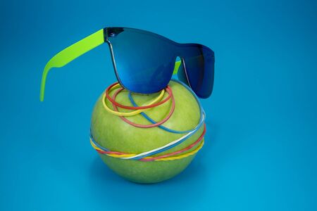 Green apple tied in colorful rubber bands and fashion sunglasses over a blue background. Simplicity back to school or diet, aerobic concept with copy spaceの写真素材