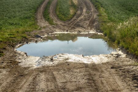 Large puddle of water on a gravel road leading through farm fields with two converging tracks on the far sideの写真素材