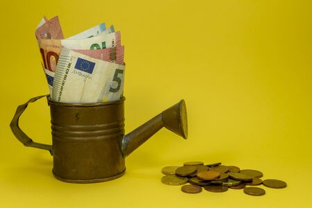 Euro banknotes of assorted denominations in a copper watering can with scattered coins on yellow with copy space for financial themesの写真素材