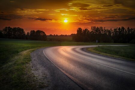 Vivid orange tropical sunset over woodland trees with a curving tarred road reflecting the color in...