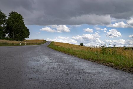 Dark grey threatening storm clouds gathering over a country road running through open fields to disappear over the hill towards blue sunny sky with white clouds in the distanceの写真素材