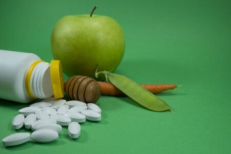 Pills spilling out of a white bottle near apple, pea pod and carrot on a green background. Healthcare, diet or weight loss conceptの写真素材