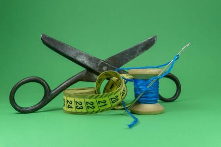 A close up image of a tape measure, old metal scissors, spool of blue thread and sewing needle on green background, handmade sewing, needlework and tailoring conceptの写真素材