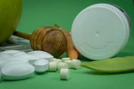 Pills spilling out of a white bottle near apple, pea pod, carrot and euro banknotes on a green background. Healthcare, diet or weight loss conceptの写真素材