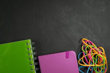 Back to school background with green spiral ring bound notebook, pink notebook and colorful rubber bands on black background. Flat lay, view from aboveの写真素材
