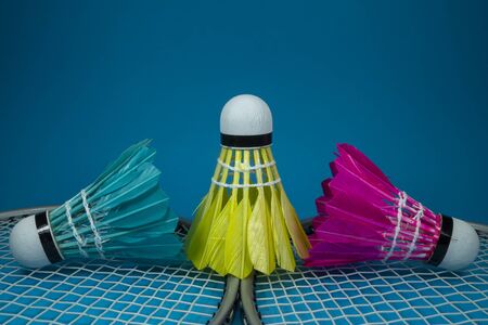 Colorful feathered shuttlecocks in blue, yellow and pink and badminton rackets on blue background in a close up viewの写真素材