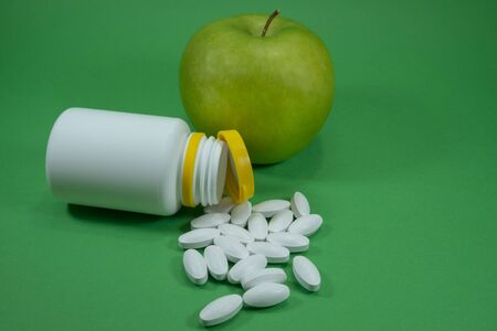 Pills spilling out of a white bottle near apple on a green background. Healthcare, diet or weight loss conceptの写真素材