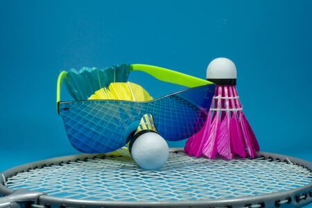 Colorful feathered badminton shuttlecock with sunglasses resting on a racket over a blue background in a close up still lifeの写真素材