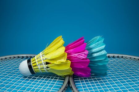 Colorful feathered shuttlecocks in blue, yellow and pink and badminton rackets on blue background in a close up viewの写真素材