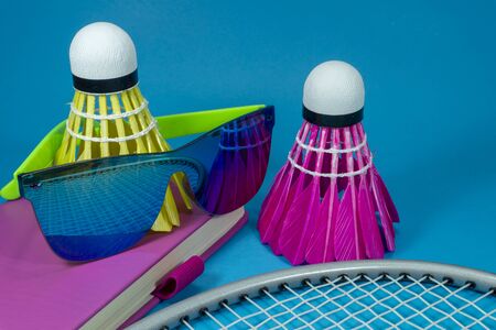 Colorful badminton shuttlecocks with racket and sunglasses resting on a brightly colored pink book or diary over a blue background in a close up still lifeの写真素材