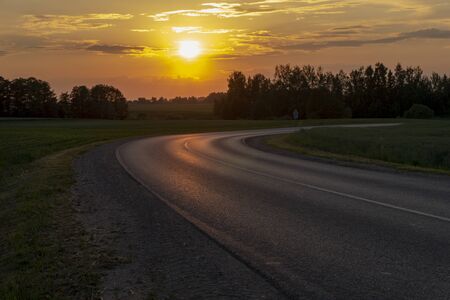Vivid orange tropical sunset over woodland trees with a curving tarred road reflecting the color in the foreground in a scenic rural landscapeの写真素材