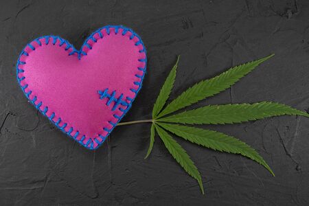 Handmade colorful felt bright pink textile heart with a fresh green marijuana leaf over a black background with copy space in a conceptual imageの写真素材