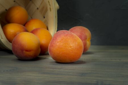 Fresh peaches spilling out of tipped wicker basket onto grey wooden tabletop. Black wall in background.の写真素材
