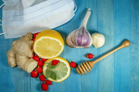Fresh healthy ingredients for a medicinal tisane or infusion with root ginger, garlic, lemon and rose hips together with a honey wand and disposable face mask to protect against airborne diseaseの写真素材