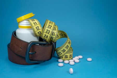 Conceptual weight loss image with a tape measure, scattered pills and genuine leather belt wound around a pharmacy bottle over blueの写真素材
