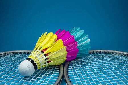 Colorful feathered shuttlecocks in blue, yellow and pink and badminton rackets on blue background in a close up viewの写真素材