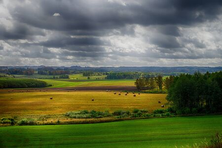 Atmospheric landscape with gathering storm clouds over agricultural fields with hay bales and woodland treesの写真素材