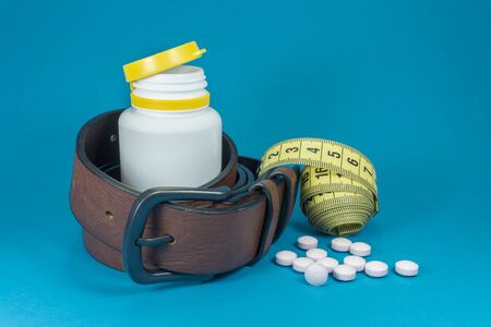 Conceptual weight loss image with a tape measure, scattered pills and genuine leather belt wound around a pharmacy bottle over blueの写真素材