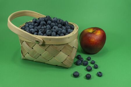 Fresh blueberries in a rustic woven basket and a red apple on a green backgroundの写真素材